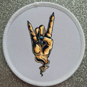 May include: Round white patch with a stitched border. The patch features a stylized illustration of a hand making the rock and roll gesture. The hand is gold with blue fingernails. The design has a flame-like element at the base.