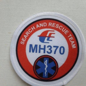 Op de afbeelding: Een witte, rode en blauwe ronde patch met de tekst "Search and Rescue Team" en "MH370" in zwart. Een blauwe ster van het leven met een slang eromheen is in het midden.