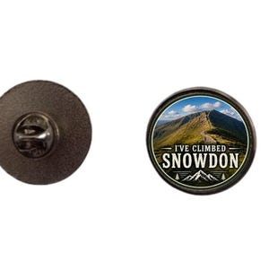 Puede incluir: Un pin redondo de esmalte con un paisaje de montaña y el texto "I'VE CLIMBED SNOWDON". El pin tiene un borde negro y un cierre metálico en la parte posterior. La imagen muestra una montaña con un camino que conduce a la cima.