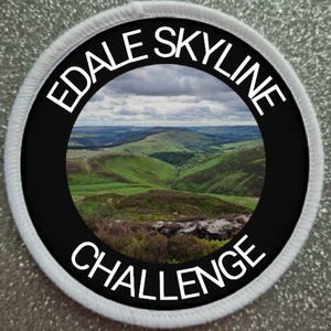 Könnte beinhalten: Runder Aufnäher mit dem Text "EDALE SKYLINE CHALLENGE" in weißen Buchstaben. Die Mitte zeigt eine Landschaft mit grünen Hügeln unter einem bewölkten Himmel, umrahmt von einem schwarzen Rand und einem weißen, genähten Rand.
