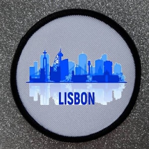 Puede incluir: Parche redondo con una silueta azul del horizonte de Lisboa sobre un fondo gris claro. La palabra "LISBON" está escrita en blanco. El parche tiene un borde negro, posiblemente un recuerdo o un emblema.