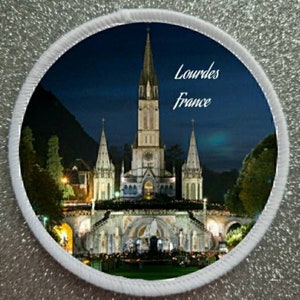 May include: A white circular patch featuring a nighttime image of the Basilica of Our Lady of the Rosary in Lourdes, France. The basilica is illuminated with lights and the text "Lourdes France" is printed in white on a dark blue background.