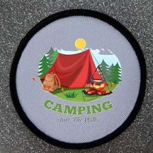 Op de afbeelding: Ronde patch met een kampeerscène. Een rode tent, rugzak en kampvuur zijn afgebeeld op een lichtgrijze achtergrond. Het woord "CAMPING" staat in het groen, met "Into The Wild" eronder. De patch heeft een zwarte rand.