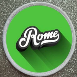 Può includere: Un cerchio bianco con un fondo verde e la parola "Roma" in un font stilizzato bianco e nero con una lunga ombra.