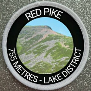 Op de afbeelding: Ronde witte patch met zwarte rand en witte tekst. De tekst luidt "Red Pike 755 Metres - Lake District". In het midden van de patch is een foto van een berg te zien.