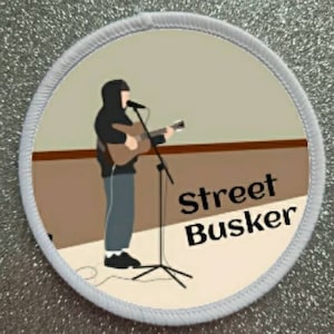 Może przedstawiać: Biała łatka z brązowym i beżowym tłem. Rysunkowa ilustracja osoby grającej na gitarze z tekstem "Street Busker" w kolorze czarnym.