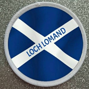 Puede incluir: Un parche redondo blanco y azul con el texto "Loch Lomand" en letras blancas sobre una franja diagonal blanca sobre un fondo azul.