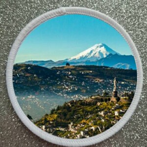May include: A white circular patch with a scenic image of a snow-capped mountain range in the background and a town in the foreground. The town is nestled in the foothills of the mountains and is surrounded by lush green vegetation.
