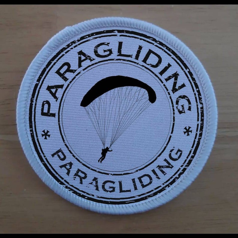 Puede incluir: Un parche redondo blanco con texto negro y una silueta negra de un parapente. El texto dice "Paragliding*Paragliding".