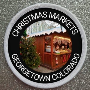 Puede incluir: Un parche circular blanco y negro con el texto "Christmas Markets Georgetown Colorado" alrededor del borde. El centro del parche presenta una foto de un puesto de mercado navideño de madera con un árbol de Navidad en primer plano.