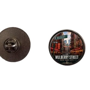 Pode incluir: Um alfinete redondo preto com uma imagem detalhada da Mulberry Street em Nova York. A imagem inclui placas de rua, edifícios e o texto "Mulberry Street - New York City."
