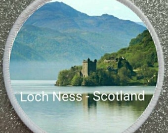 May include: A white round patch with a photo of a castle on a small island in a lake. The lake is surrounded by mountains and trees. The text "Loch Ness - Scotland" is printed below the image.