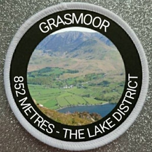 May include: White round patch with a black circle in the center. The black circle has white text that reads "Grasmoor 852 Metres - The Lake District". Inside the black circle is a photo of a mountain range and a lake.