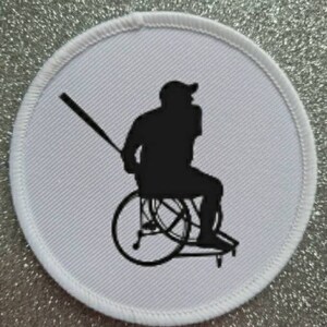 May include: White round patch with a black silhouette of a person in a wheelchair swinging a baseball bat.