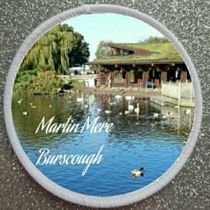 Puede incluir: Un parche redondo blanco con una foto de un lago con un edificio de madera y un techo verde. El texto "Martin Mere Burscough" está impreso en el parche.
