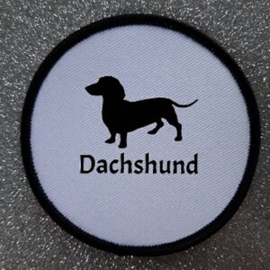 Puede incluir: Parche redondo blanco con una silueta negra de un dachshund y la palabra "Dachshund" en texto negro.