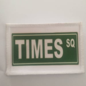 Könnte beinhalten: Weißer Stoff-Patch mit grünem Rand und dem Text "TIMES SQ" in Weiß.