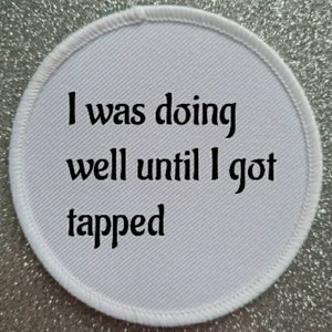 Könnte beinhalten: Ein runder, weißer Aufnäher mit dem schwarzen Text "I was doing well until I got tapped". Er hat einen weißen, genähten Rand und liegt auf einem glitzernden, silbernen Hintergrund.