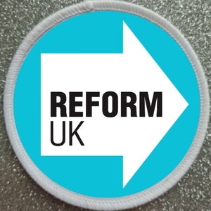 Op de afbeelding: Een witte ronde patch met een witte pijl die naar rechts wijst. De pijl bevat de tekst "REFORM UK" in zwart.