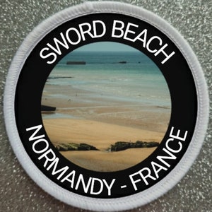 May include: Round patch with a beach scene, featuring the text "SWORD BEACH NORMANDY - FRANCE" in white lettering on a black background, surrounded by a white border. The image inside shows a sandy beach and the ocean.