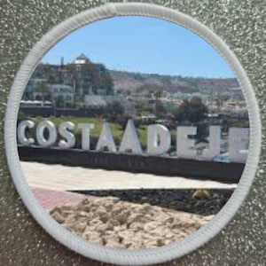Puede incluir: Un parche circular blanco con una foto de un cartel que dice "Costa Adeje" en letras blancas sobre un fondo de césped verde. El cartel está frente a un edificio y árboles. La foto está tomada desde un ángulo bajo.