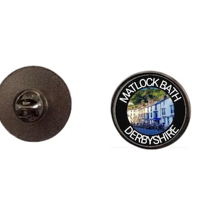 May include: A circular pin badge with a dark metal finish. The badge displays a scenic image of buildings and the text "MATLOCK BATH DERBYSHIRE" in a black border.