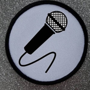 May include: A white round patch with a black outline. The patch features a black microphone with a white grid pattern on the microphone head. The microphone is angled and the cord is curved.