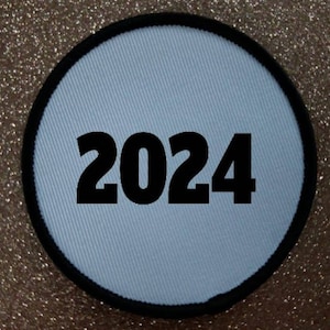 Puede incluir: Un parche redondo blanco con un borde negro y el año 2024 impreso en negro.