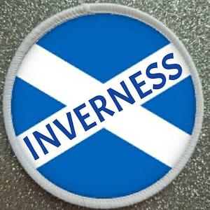 May include: A white round patch with a blue and white Scottish flag design. The word "INVERNESS" is written in blue letters across the flag.