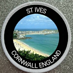 May include: A white round patch with a black circle in the center. The black circle has a white border and a picture of a beach scene with a town in the distance. The text "ST IVES CORNWALL ENGLAND" is written in white around the black circle.