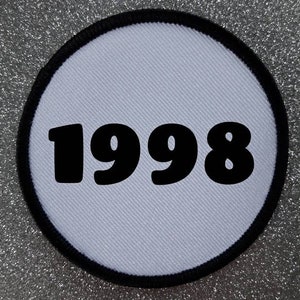 Könnte beinhalten: Runder weißer Patch mit schwarzem Rand und der Jahreszahl 1998 in Schwarz gedruckt.