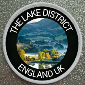 May include: White round patch with black border and text "THE LAKE DISTRICT ENGLAND UK". The centre of the patch features a scenic image of a lake and hills.