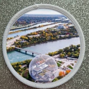 May include: A white circular patch featuring a photograph of a large geodesic dome structure surrounded by a river and trees. The dome is a well-known landmark in Montreal, Canada.