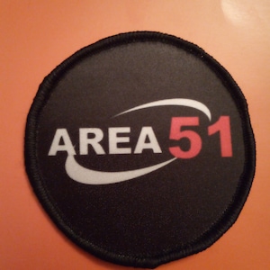 Könnte beinhalten: Ein runder schwarzer und weißer Patch mit dem Text "AREA 51" in roten und weißen Buchstaben. Der Text befindet sich in einem gekrümmten weißen Bogen mit einem weißen Schwung darunter.