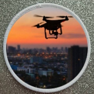 Könnte beinhalten: Eine schwarze Drohne fliegt bei Sonnenuntergang über eine Stadtkulisse. Die Drohne hat vier Propeller und eine Kamera, die an der Unterseite befestigt ist. Die Stadt ist mit Lichtern beleuchtet, und der Himmel ist in leuchtenden Orange- und Rosatönen.