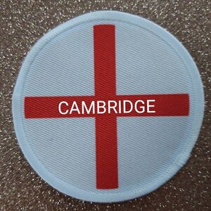 Op de afbeelding: Een witte ronde patch met een rood kruis in het midden. De woorden "CAMBRIDGE" zijn in zwarte letters over het midden van het kruis gedrukt.