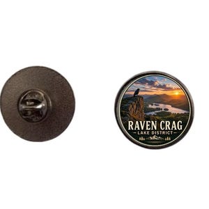 May include: A round pin with a black border and a scenic image of a raven perched on a cliff overlooking a lake at sunset. The text "RAVEN CRAG - LAKE DISTRICT" is below the image. The pin's back is dark gray.