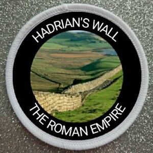 Könnte beinhalten: Ein weißer und schwarzer runder Patch mit dem Text "Hadrian's Wall The Roman Empire" am Rand. Die Mitte des Patches zeigt ein Foto des Hadrianswalls in der Landschaft.