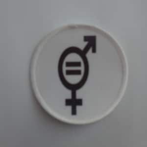 Può includere: Un cerotto rotondo bianco con un simbolo nero di uguaglianza di genere. Il simbolo è una combinazione dei simboli femminile e maschile con un segno uguale al centro.