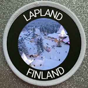 Puede incluir: Un parche redondo blanco con un círculo negro en el centro. El círculo negro tiene las palabras "LAPLAND FINLAND" en texto blanco. Dentro del círculo negro hay una foto de un paisaje nevado.