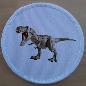 Puede incluir: Un parche redondo blanco con un dinosaurio marrón y verde con la boca abierta. El dinosaurio es un Tyrannosaurus Rex.