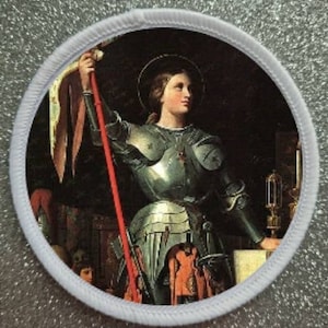 May include: A white embroidered patch with a colorful image of a female figure in armor holding a spear. The figure is wearing a helmet and a red cape. The background is dark and includes a candle and a book.