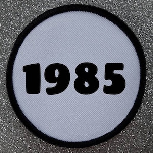 Peut inclure: Patch rond blanc avec une bordure noire et le nombre 1985 imprimé en noir.