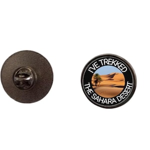 May include: A circular black enamel pin badge. The design features a desert scene with palm trees and sand dunes, and the text "I've trekked the Sahara Desert".
