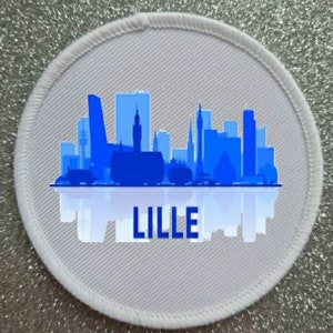 Puede incluir: Parche redondo blanco con una silueta azul del horizonte de Lille y la palabra "LILLE" en texto azul.