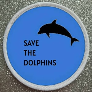 May include: A white circular patch with a blue background and a black silhouette of a dolphin jumping out of the water. The text "SAVE THE DOLPHINS" is printed in black below the dolphin.