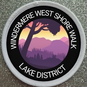 May include: A white circular patch with black text and a silhouette of a mountain range with a tree in the foreground. The text reads "Windermere West Shore Walk Lake District".