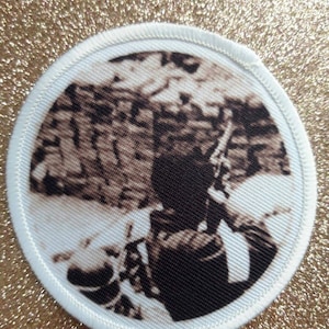 May include: Circular patch featuring a sepia-toned image of a person. The patch has a white border with stitched edges. The image appears to be a photograph or illustration. The background is a glittery gold colour.