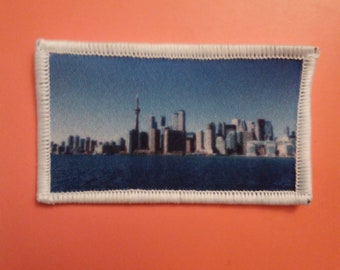 May include: A blue and white embroidered patch featuring a cityscape skyline with a tall building in the center.