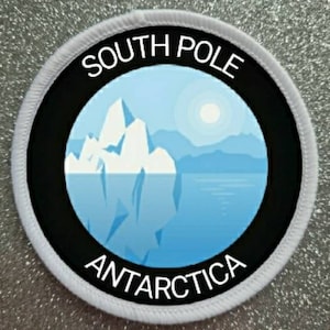 Könnte beinhalten: Ein weißer runder Patch mit schwarzem Rand und einer blauen und weißen Illustration eines Eisbergs vor einer Bergkette. Der Text "SOUTH POLE" ist oben auf dem Patch in Schwarz und "ANTARCTICA" ist unten in Schwarz.